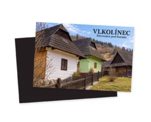 Grafická magnetka zobrazujúca bielu, zelenú a tmavú drevenicu so strmými strechami, plotom a konármi stromov s nápisom Vlkolínec – Drevenice pod horami.